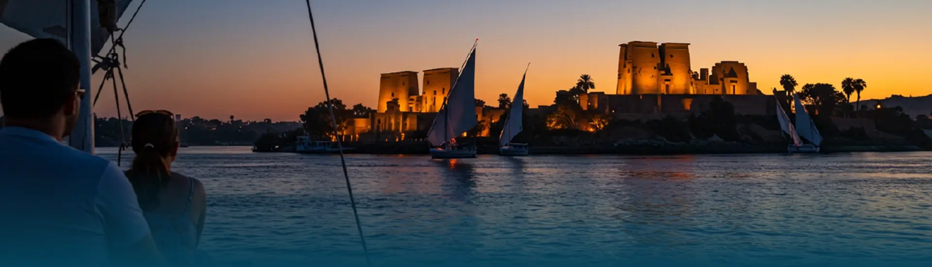 Sunset view of an illuminated Egyptian temple during a Nile cruise from Luxor to Aswan with feluccas.