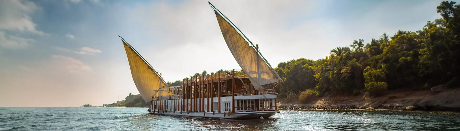 Traditional Dahabiya Nile cruise boat with large sails sailing past green riverbanks on the Nile River in Egypt.