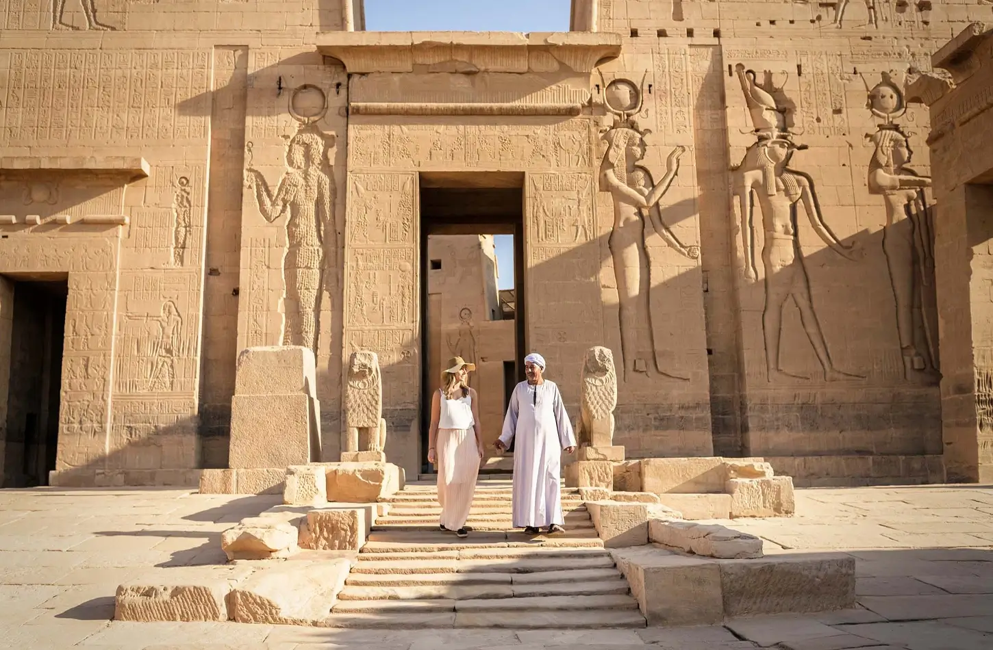 A man and woman walk down stone steps in front of a massive ancient Egyptian temple wall with hieroglyph carvings.