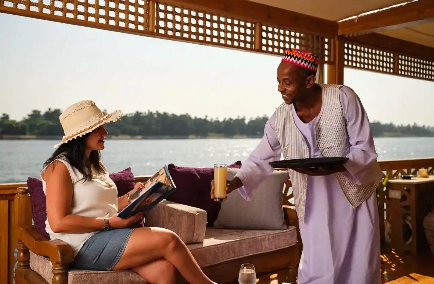 A server in a white galabeya hands a drink to a seated woman wearing a sun hat on a boat deck