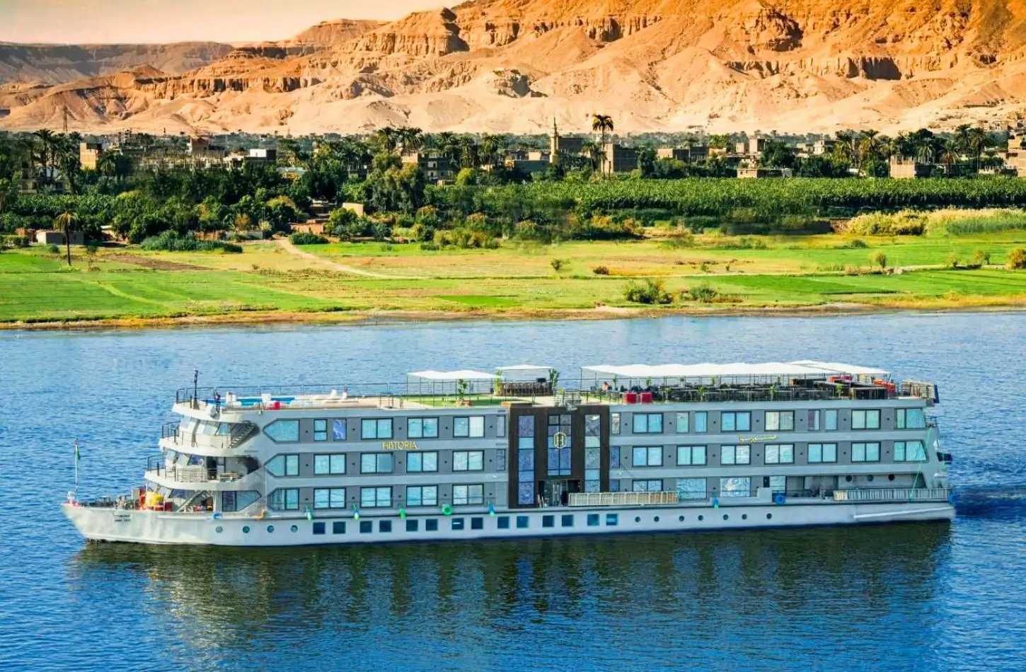 The luxury cruise ship Historia sailing along the Nile River past lush green banks and desert mountains, demonstrating is it safe to go on a Nile cruise.
