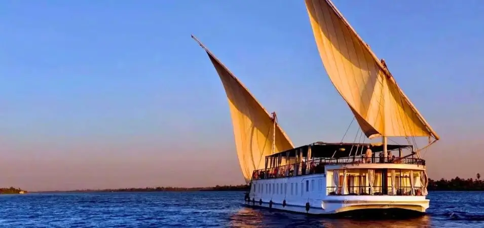 Nile Cruise Cost 2026: Prices by Ship Type & Route 2 Traditional wooden twin-sail Dahabiya sailboat cruising along the calm waters of the Nile River under a clear blue sky, illustrating the experience and luxury involved in calculating the Nile Cruise Cost.