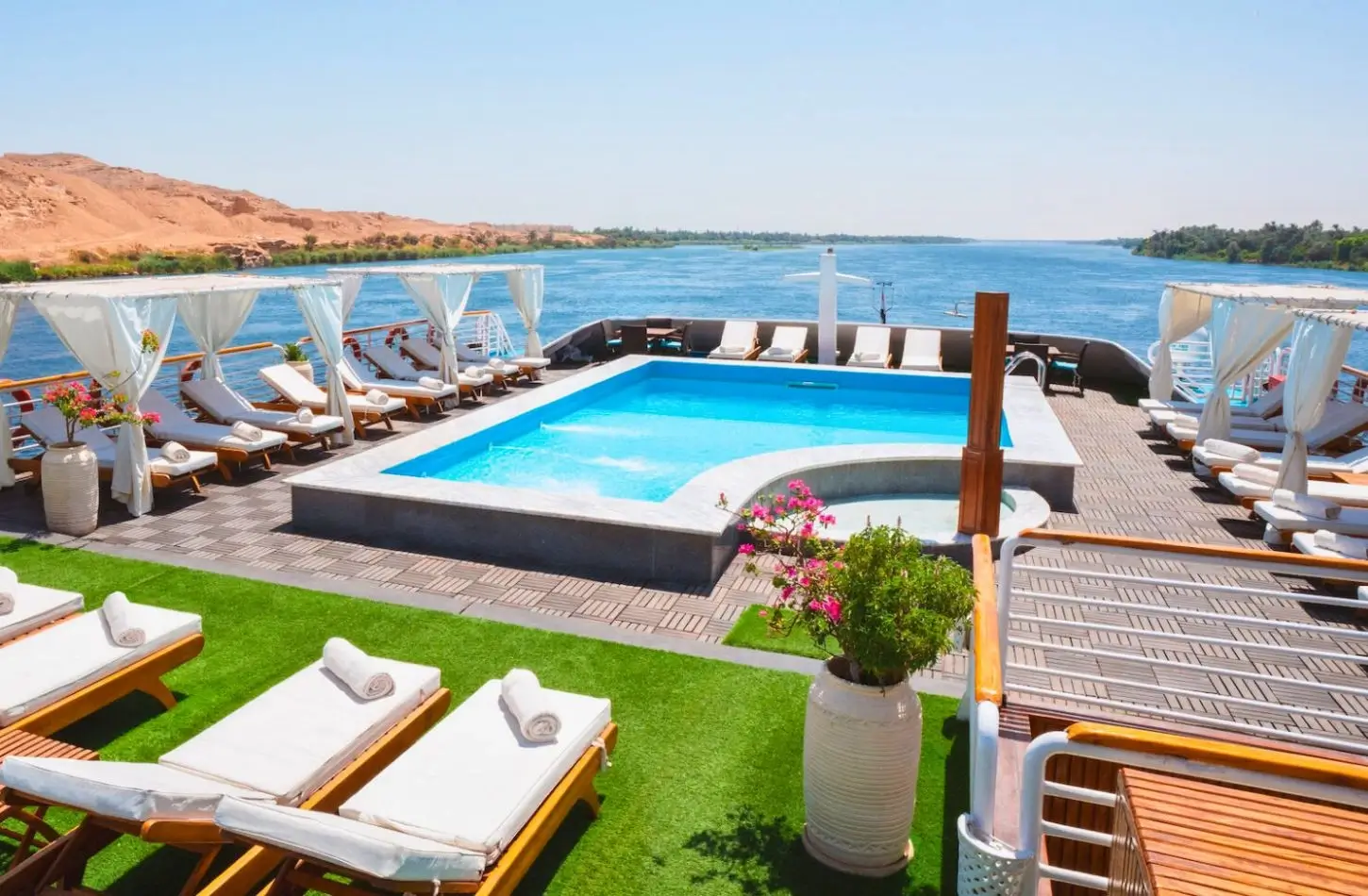 The top sun deck of a luxury riverboat featuring a swimming pool, shaded cabanas, and lounge chairs cruising down the blue Nile River, illustrating is it safe to go on a Nile cruise.