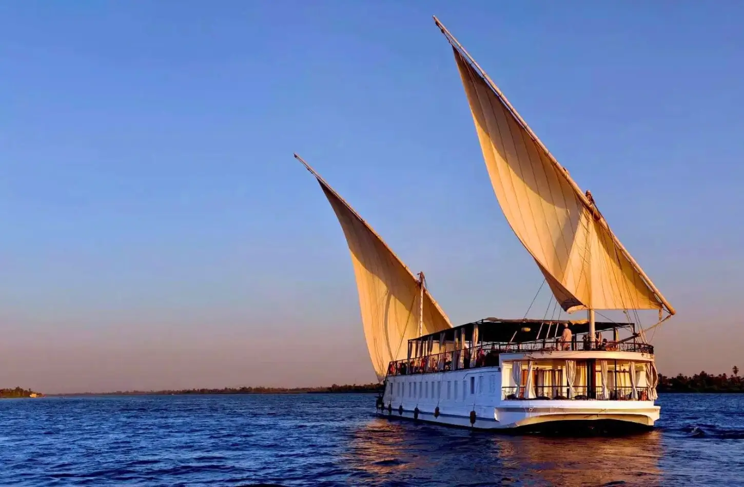 A white wooden Dahabiya with large dual sails cruising down the Nile River, showcasing luxury boutique options for a Nile Cruise Cost comparison.